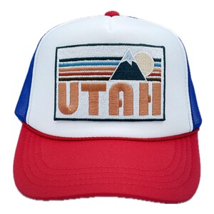 May include: A white and red trucker hat with a blue mesh back. The front of the hat features an embroidered patch with a mountain range, a sun, and the word "UTAH" in a retro style.