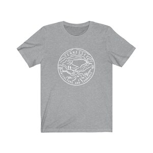 May include: A gray t-shirt with a white graphic of the Tennessee state seal. The seal features a mountain landscape with a river and the words "Tennessee, Agriculture and Commerce".