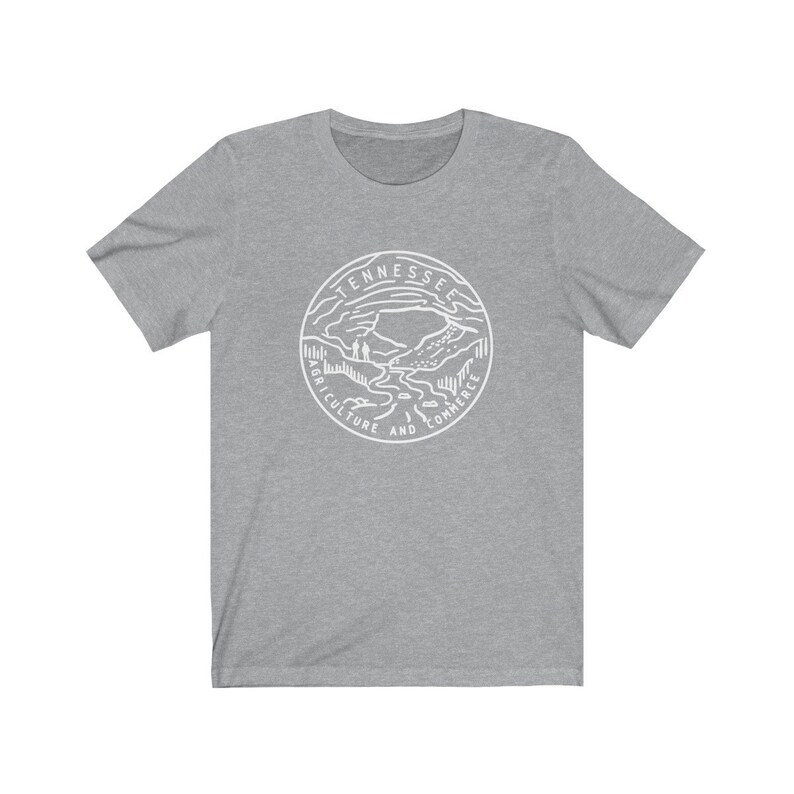May include: A gray t-shirt with a white graphic of the Tennessee state seal. The seal features a mountain landscape with a river and the words "Tennessee, Agriculture and Commerce".