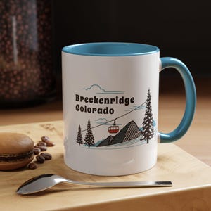 May include: White ceramic mug with a blue rim and handle. The mug features a graphic of a mountain scene with a ski lift and the text "Breckenridge Colorado".