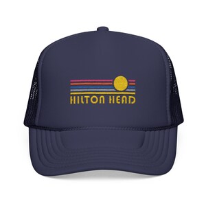 May include: A navy blue trucker hat with a mesh back. The front of the hat features an embroidered design of a sunset with a yellow sun and the text "HILTON HEAD" in yellow.