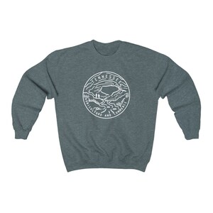 May include: A dark heather gray sweatshirt with a white graphic of the state seal of Tennessee. The seal features a mountain range, a river, and the words "Agriculture and Commerce".
