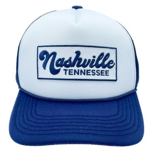 May include: A white trucker hat with a blue mesh back. The front of the hat has a blue patch with the text "Nashville Tennessee" in a cursive font.
