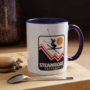 May include: White ceramic mug with a blue rim and handle. The mug features a retro-style illustration of a skier in the mountains with the text "Steamboat Colorado".