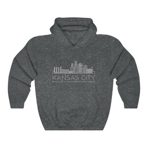 Może przedstawiać: Ciemnoszara bluza z kapturem z białym rysunkiem linii horyzontu Kansas City. Tekst "KANSAS CITY" jest wydrukowany na biało pod linią horyzontu. Bluza ma kaptur ze sznurkiem i kieszeń kangura.