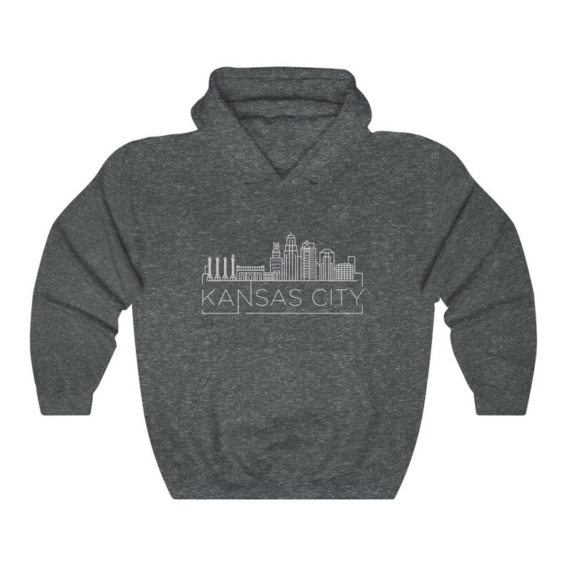 Może przedstawiać: Ciemnoszara bluza z kapturem z białym rysunkiem linii horyzontu Kansas City. Tekst "KANSAS CITY" jest wydrukowany na biało pod linią horyzontu. Bluza ma kaptur ze sznurkiem i kieszeń kangura.