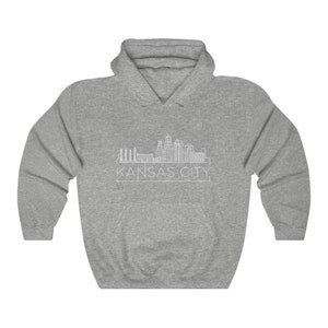 Może przedstawiać: Szara bluza z kapturem z białym rysunkiem linii horyzontu Kansas City. Nazwa miasta, 'KANSAS CITY', jest wydrukowana poniżej na biało. Bluza ma przednią kieszeń i kaptur ze sznurkiem.