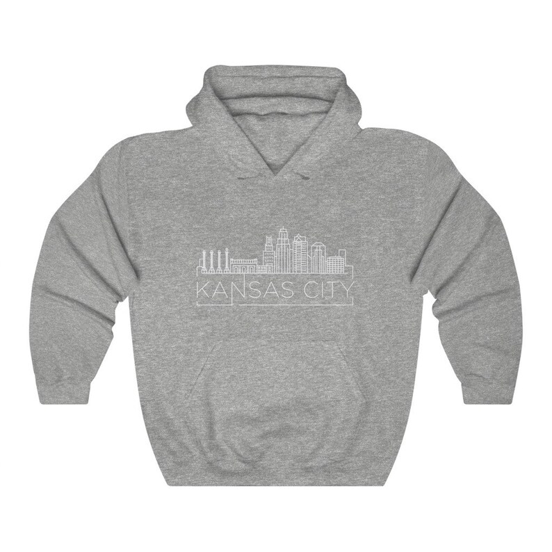 Może przedstawiać: Szara bluza z kapturem z białym rysunkiem linii horyzontu Kansas City. Nazwa miasta, 'KANSAS CITY', jest wydrukowana poniżej na biało. Bluza ma przednią kieszeń i kaptur ze sznurkiem.
