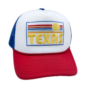 May include: A white trucker hat with a red bill. The front of the hat features a blue and yellow embroidered patch with the word "TEXAS" and a stylized sunset design.