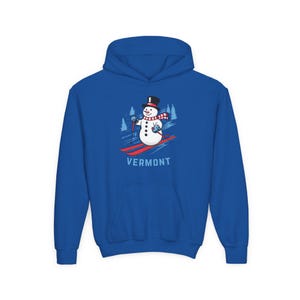 May include: A royal blue hoodie featuring a graphic of a snowman skiing down a slope. The snowman wears a black top hat, a red and white scarf, and holds ski poles. The word "VERMONT" is printed below the skier.