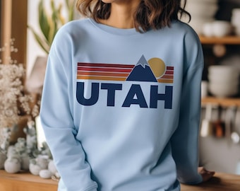 May include: A light blue sweatshirt featuring a retro-style graphic of a mountain range, a sun, and the word "UTAH" in navy blue.