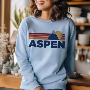 May include: A light blue sweatshirt with a retro-style graphic of a mountain, sun, and the word "ASPEN" in navy blue.