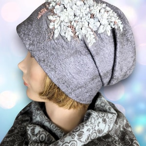May include: A gray beanie-style hat with a floral design. The hat features a cluster of white and light blue flowers with delicate leaves and accents. The hat is paired with a patterned scarf, creating a coordinated look. The hat is made of a soft, textured material.