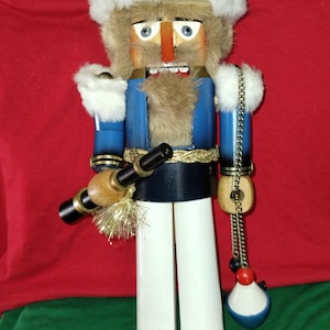 May include: A wooden nutcracker figurine in a fisherman theme. It features a white fur hat, blue jacket, white pants, and a fishing rod. The nutcracker holds a chain with a bobber and a red and black bead.