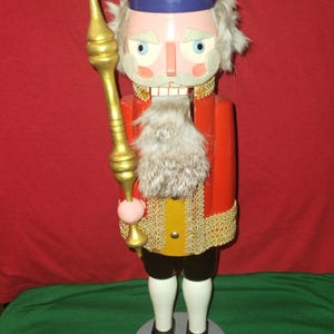 Könnte beinhalten: Eine Holz-Nussknacker-Figur in königlicher Kleidung. Sie hat eine goldene Krone, eine rote Jacke mit goldenen Verzierungen und einen Kunstpelzbart. Der Nussknacker hält einen goldenen Stab und steht auf einem grauen, kreisförmigen Sockel.