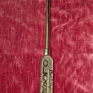 May include: A gold-toned metal hook tool featuring a long, slender shaft and a rectangular handle. The handle has a textured, cross-hatched surface with the word CLOSED embossed vertically in capital letters, providing a clear grip for the user.