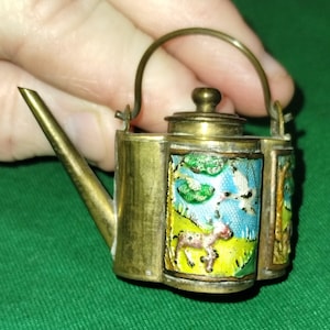 Puede incluir: Tetera de latón en miniatura con tapa abisagrada y asa curva. Los lados presentan escenas de esmalte colorido de un ciervo en un campo, árboles y pájaros contra un cielo azul. El pico es largo y delgado.