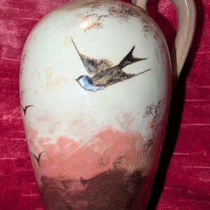 May include: A ceramic jug with a glossy, crackled glaze finish. The body features a hand-painted scene of a flying swallow against a cream-colored background, transitioning into a soft pink midsection and a dark brown base with silhouettes of birds.
