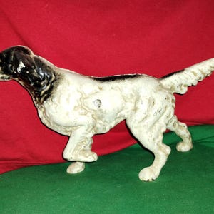 Può includere: Statuetta di cane in ghisa in posizione di camminata. Il cane è principalmente bianco con macchie nere sulla testa e sulla schiena. La statuetta ha una superficie strutturata ed è posta su uno sfondo rosso e verde.