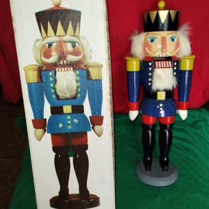 Puede incluir: Una figura de cascanueces de madera con una chaqueta azul, botas negras y una corona dorada está junto a su caja. La caja presenta una ilustración a juego. El cascanueces tiene barba y bigote blancos y está sobre una base gris.