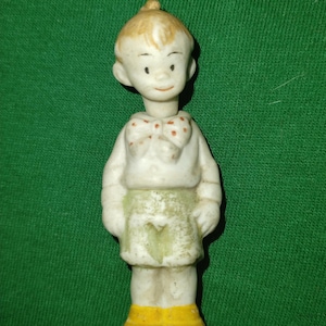 以下が含まれることがあります： 子供の小さなヴィンテージ磁器フィギュア。フィギュアは肌の色が明るく、ブロンドの髪で、赤い水玉模様の蝶ネクタイが付いた白いシャツ、緑色のスカート、黄色のブーツを着用しています。フィギュアは直立しています。