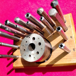 May include: A collection of metal punches and tools, including cylindrical and hexagonal shapes, set in a wooden holder. The tools vary in size and have hollow tips. Numbers 8, 9, and 15 are visible on the wooden base.