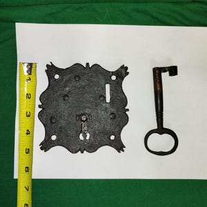 Puede incluir: Una placa de cerradura antigua de metal negro ornamentada con una llave a juego. La placa de la cerradura mide aproximadamente 10 cm de ancho y tiene una forma decorativa e irregular. La llave es de color marrón oscuro y tiene un mango simple y redondo. Los artículos están sobre una superficie blanca.