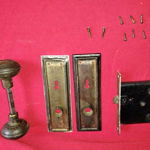 Puede incluir: Juego de herrajes de puerta antiguos que incluye un pomo de puerta de metal oscuro, dos placas rectangulares con cerraduras, un pestillo de puerta negro y varios tornillos pequeños. El hardware tiene un aspecto vintage y envejecido.