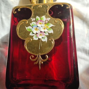 Puede incluir: Frasco de perfume rectangular de vidrio rojo rubí con tapa y detalles de metal dorado. La parte delantera presenta un panel decorativo dorado con un diseño floral pintado a mano en blanco, rosa, azul y amarillo.