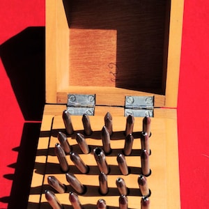 May include: Wooden box with a hinged lid, containing a grid of metal punches. The box is open, revealing the tools arranged in rows. The background is a solid red color.