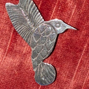 May include: A silver-toned hummingbird brooch featuring a polished metallic finish. The bird's body is decorated with a repeating pattern of small, embossed heart shapes, while the wings and tail display fine, etched linear feather details.