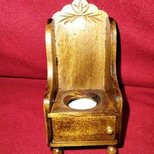 May include: A small, wooden candle holder shaped like a chair. The candle holder is stained a warm brown color and features a decorative carved design on the back. It has a small drawer and rests on four rounded feet.