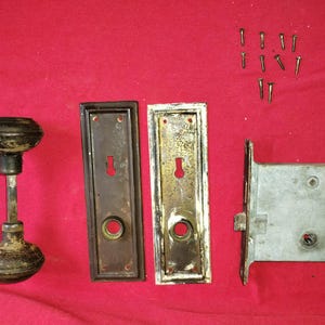Può includere: Set di ferramenta per porte antiche, tra cui una maniglia nera e bronzo, due placche rettangolari, un chiavistello in metallo e diverse viti. L'hardware è disposto su una superficie rossa.