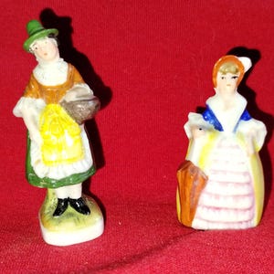 Puede incluir: Dos pequeñas figuras de cerámica de mujeres con vestimenta histórica. Una figura lleva un sombrero verde, una parte superior naranja, un delantal amarillo y sostiene una cesta. La otra figura viste un vestido azul y blanco con un abrigo amarillo y un accesorio marrón.