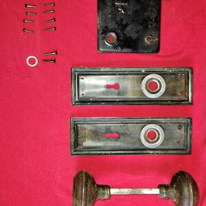 May include: An antique door hardware set, including a black lock, two rectangular plates with keyholes, and a pair of brown doorknobs connected by a metal rod. Several screws and a white washer are also present, all against a red background.