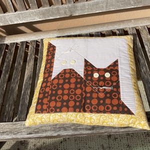 Könnte beinhalten: Quadratisches Zierkissen mit einem Katzendesign. Der Körper der Katze ist braun mit orangefarbenen Punkten, und der Kopf ist einfarbig braun. Der Hintergrund ist weiß mit grauen Streifen. Das Kissen hat einen gelben Rand mit einem floralen Muster.