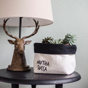 May include: A white fabric planter with a black top rim and the text "Mutha Succa" printed on the front. The planter is filled with green succulents and is sitting on a black side table next to a lamp with a deer head base.