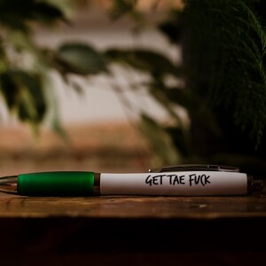 Puede incluir: Un bol&iacute;grafo blanco con un cuerpo verde y el texto "GET THE FUCK" escrito en &eacute;l.