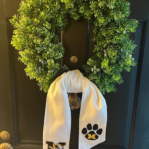 May include: A green, leafy wreath with a white sash hanging below. The sash features the text "Mizzou" and a paw print logo with the letter "M" in the center. The wreath is hanging on a black door.