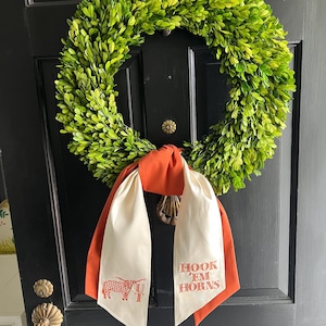 May include: A vibrant green boxwood wreath hangs on a black door, adorned with a burnt orange and cream-colored scarf. The scarf features the text "HOOK 'EM HORNS" and a longhorn illustration in red.