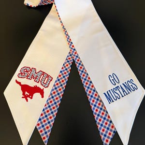 May include: A white graduation stole featuring a red embroidered SMU logo with a mustang silhouette on one side and the blue embroidered text GO MUSTANGS on the other. The inner edges are lined with a red, white, and blue checkered pattern.