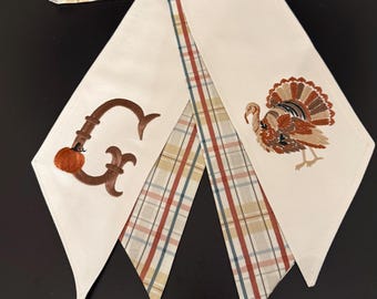 May include: A cream-colored table runner featuring a brown embroidered letter G with a small orange pumpkin, and a detailed embroidered turkey. The runner has a pointed end design and a reverse side with a plaid pattern in blue, tan, and red.