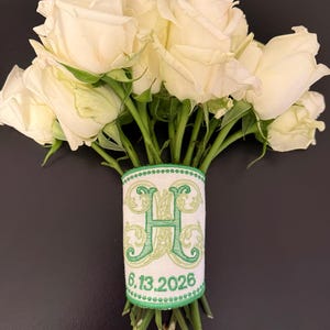 May include: A bouquet of white roses with green stems wrapped in a white fabric ribbon featuring a large embroidered green letter H and the date 6.13.2026 in green thread, finished with a green stitched border along the top and bottom edges.
