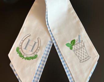 May include: White sash with blue gingham trim. One end features a horseshoe and the letter "H" embroidered in blue, surrounded by a green wreath. The other end has an embroidered cocktail with a mint sprig and a blue straw.