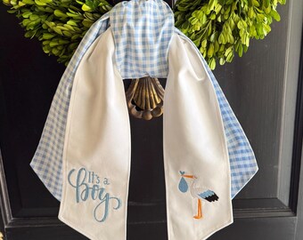 May include: A decorative fabric door hanger featuring a light blue and white gingham pattern. The white fabric tails are embroidered with the blue text It's a Boy and a stork carrying a blue bundle. The hanger is draped over a green leaf wreath.