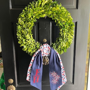 May include: A green boxwood wreath hangs on a black door, adorned with a navy blue scarf featuring red and white stars and embroidered fireworks. The scarf also has the embroidered letters "F" and "H".