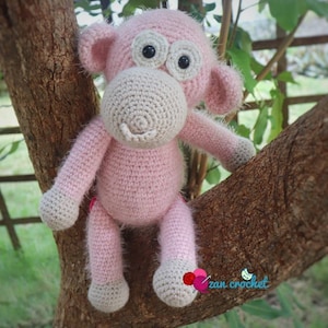 May include: A pink crocheted monkey toy with brown eyes and a brown nose, sitting on a tree branch. The monkey has a fluffy texture and is holding onto the branch with its hands.