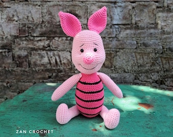 May include: A pink crocheted pig with black eyes and a black and pink striped shirt. The pig is sitting on a green surface.