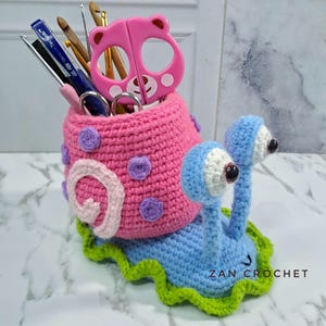 Puede incluir: Un organizador de escritorio en forma de caracol tejido a mano en rosa y azul. La concha del caracol es rosa con lunares morados, y el cuerpo es azul con un borde verde. Contiene bolígrafos, tijeras y ganchillos. El texto "ZAN CROCHET" es visible.