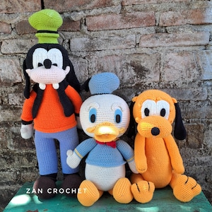 Puede incluir: Tres muñecos de personajes de Disney hechos a mano con ganchillo: Goofy, el Pato Donald y Pluto. Goofy lleva una camisa naranja, pantalones azules y un sombrero verde. El Pato Donald viste camisa y gorro azules con pajarita roja. Pluto es naranja con orejas negras.